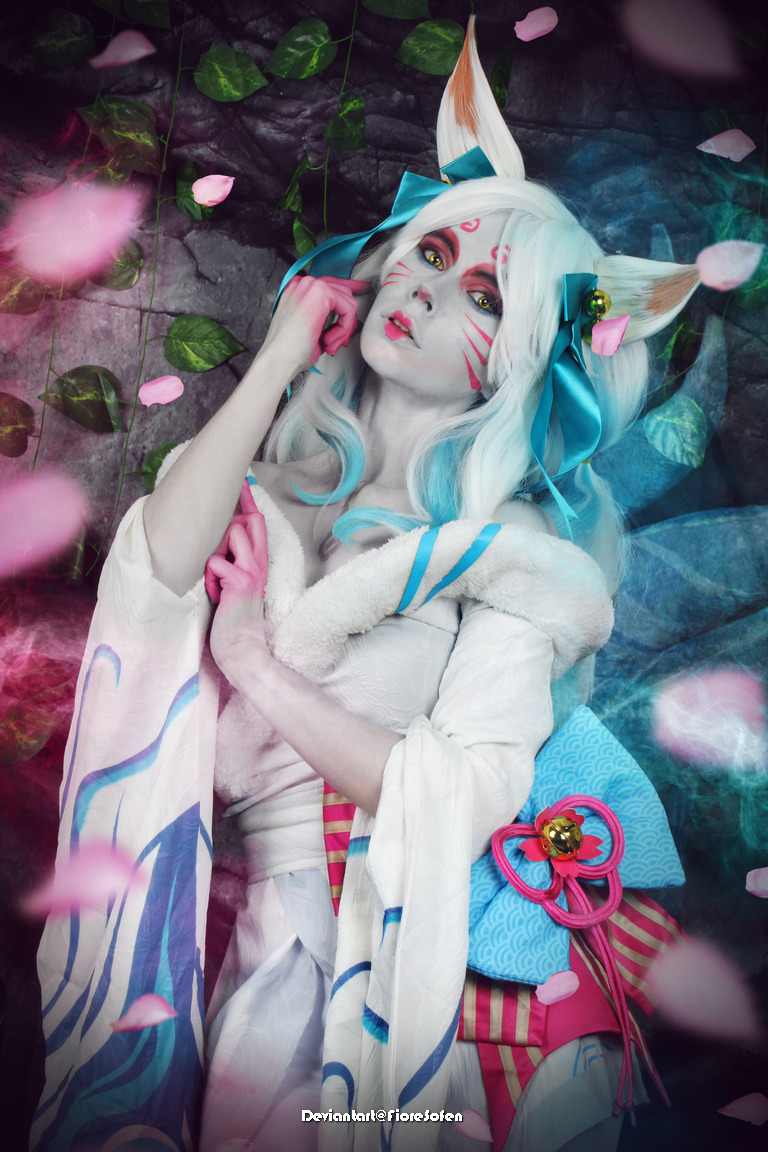 A cosplayer dressed as a character with fox-like features, featuring white hair, colorful ribbons, and a flowing garment. The background includes green leaves and pink petals, creating a mystical atmosphere.