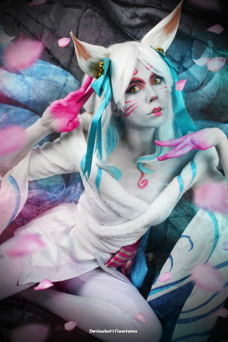 A person in costume representing a fantasy character with fox-like features, including ears and painted facial patterns, adorned with blue ribbons and surrounded by pink petals.