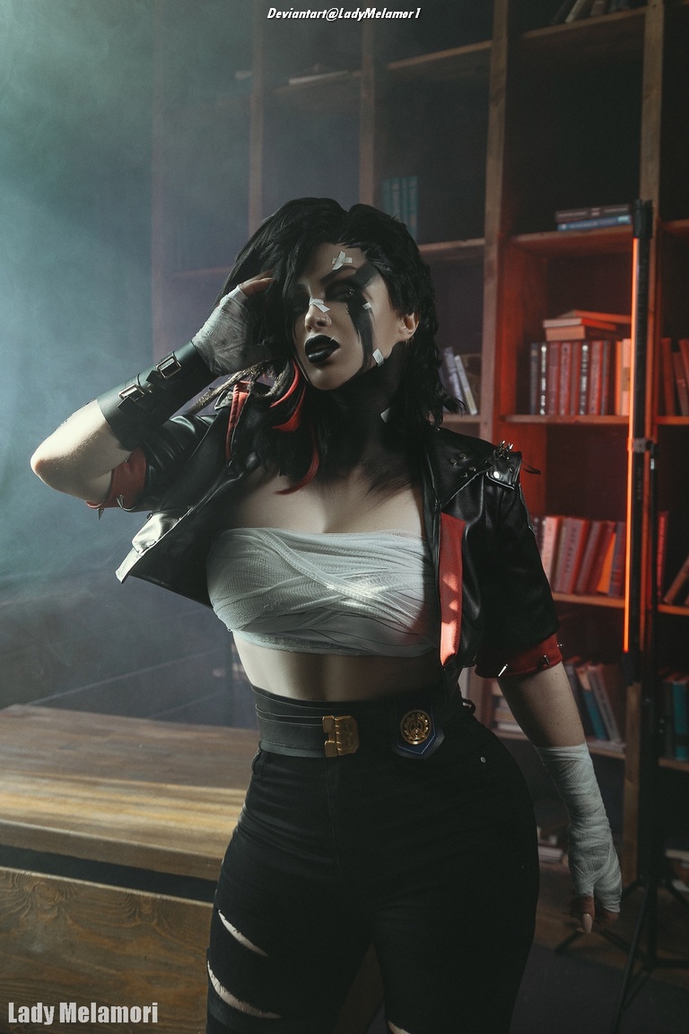 A person with dramatic makeup and styled black hair poses in a smoky room, wearing a cropped top and a leather jacket, with a bookshelf in the background.