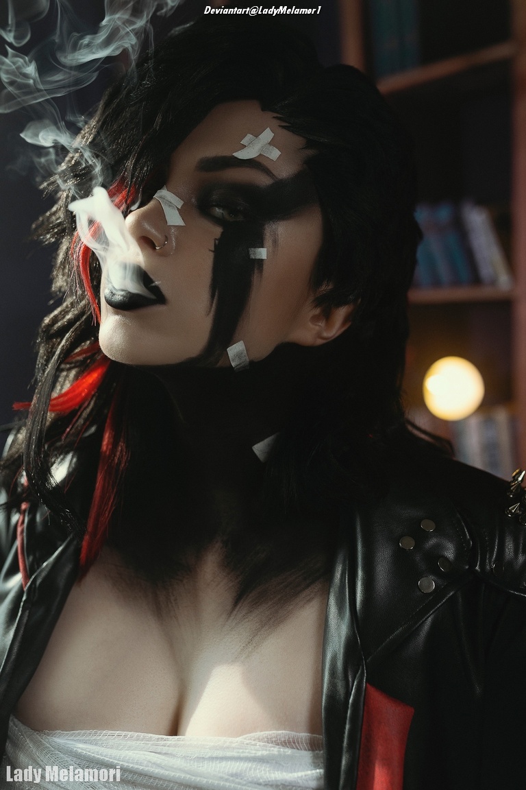 A close-up portrait of a person with striking makeup featuring black and red colors, looking contemplative while exhaling smoke. They wear a black leather jacket adorned with studs and have pieces of tape on their face.