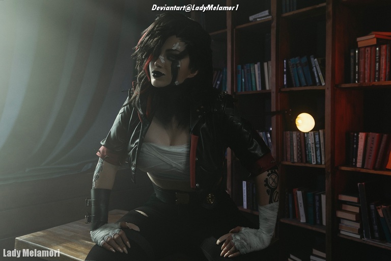 A person dressed in a punk-style outfit with dark hair and makeup, sitting in a well-lit room filled with bookshelves. The individual has a confident expression and wears a black jacket with red accents.