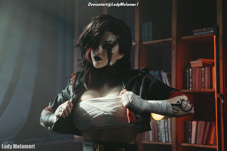 A cosplayer portraying a character with dark hair and striking makeup, wearing a black leather jacket and bandages around the torso, standing in a room with bookshelves.