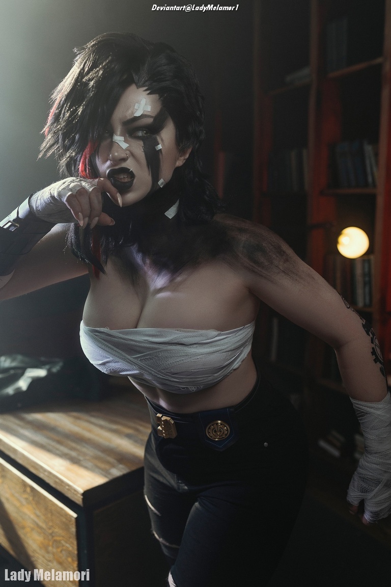 A person posing dramatically in a dark room with bookshelves, wearing a partially wrapped top and black pants, with bold makeup and a fierce expression.