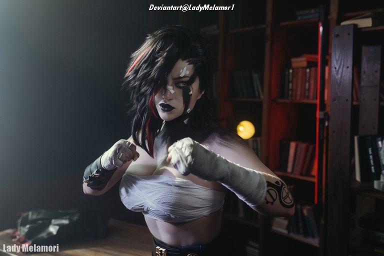 A person with a dramatic hairstyle and makeup, posing fiercely in a dimly lit room with bookshelves in the background.