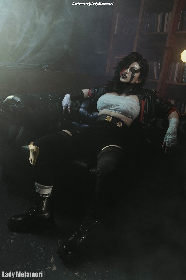 A person dressed in a black leather jacket with a cropped top and combat boots, sitting on a black couch, surrounded by smoke and bookshelves.