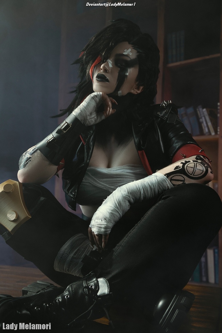 A person with dark hair and striking makeup, dressed in an edgy black leather outfit, sitting on the floor with a confident expression. The setting includes bookshelves in the background, suggesting an indoor environment.