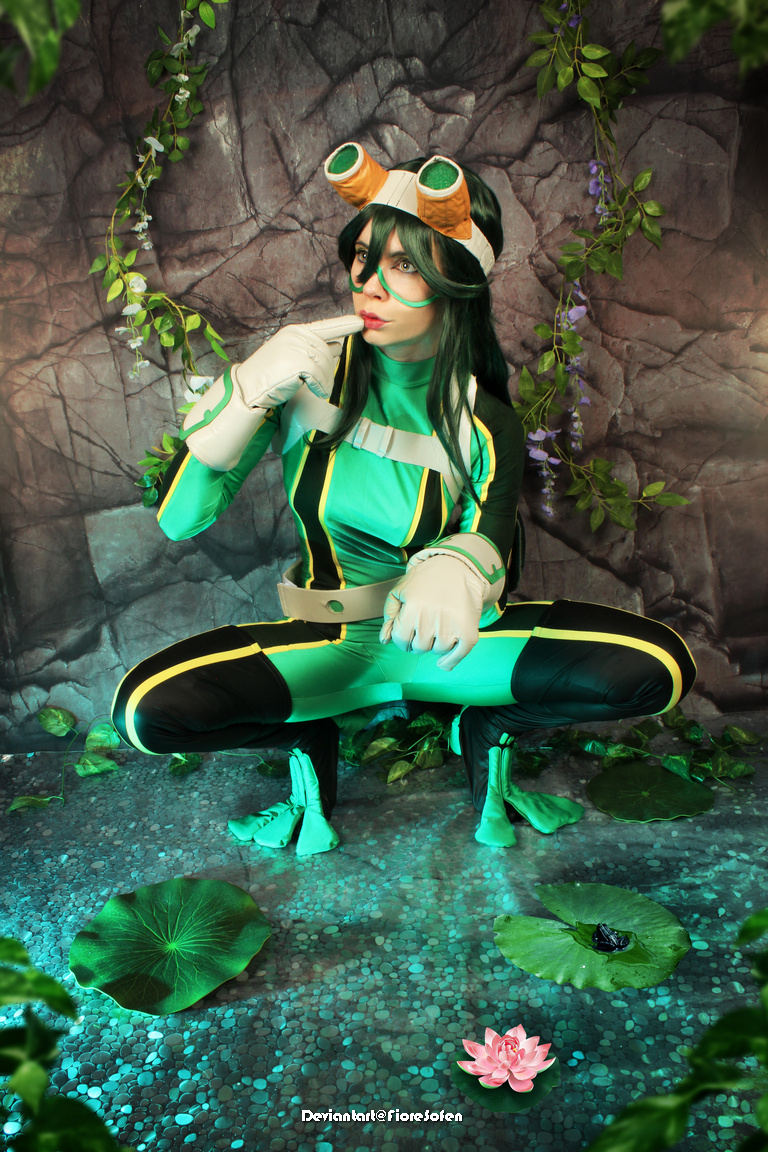 A cosplayer dressed as a frog-themed character, squatting near water lilies in a lush, green setting with a rocky background.