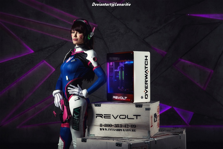 Cosplayer dressed as D.Va from Overwatch, standing confidently next to a computer setup, with purple geometric background.