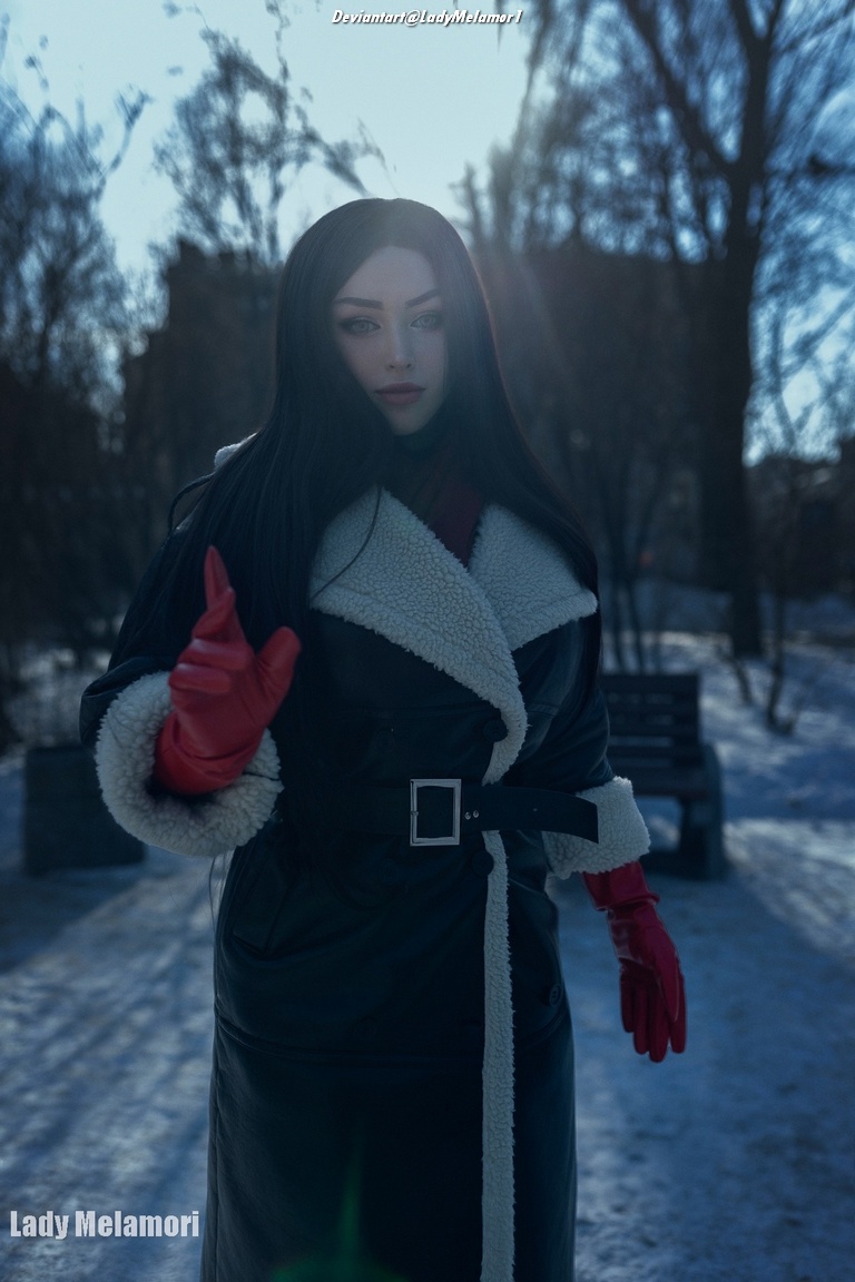 A person with long black hair wearing a black coat with a white fur collar and red gloves, standing outdoors in a snowy landscape.