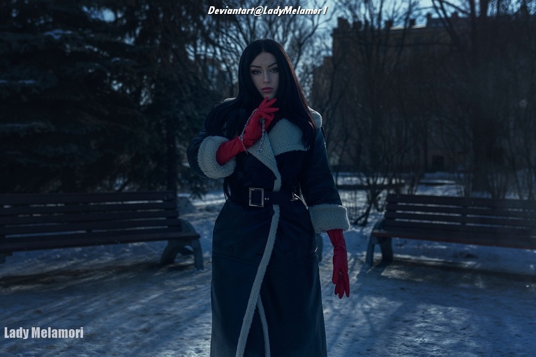 A woman with long dark hair wearing a blue coat with a fur collar and red gloves, standing in a snowy outdoor setting with benches in the background.
