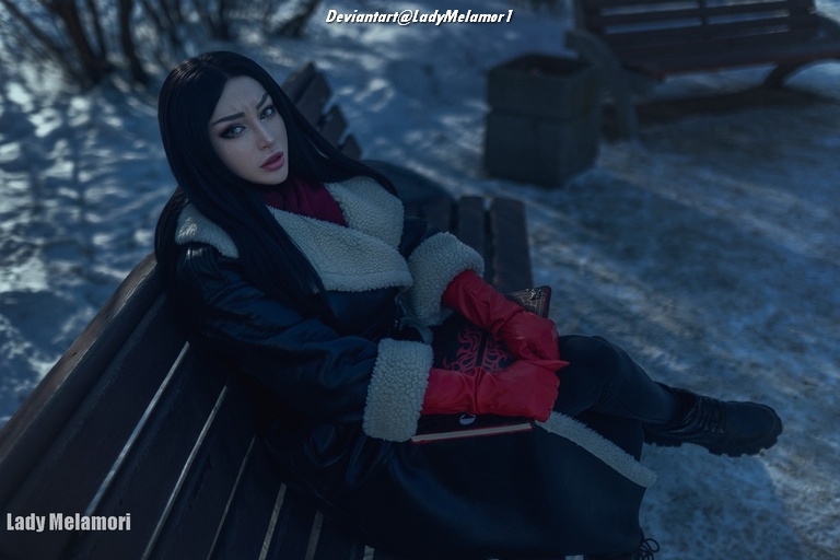 A person with long black hair wearing a black coat with a fur collar and red gloves, sitting on a bench in a snowy outdoor setting, holding a book.