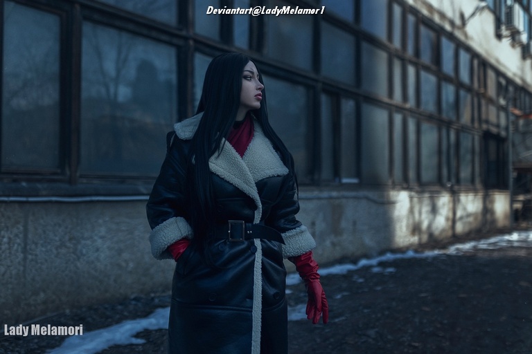A person with long black hair stands in an urban setting, wearing a black coat with a fur collar and red gloves, looking thoughtfully into the distance next to a dilapidated building.