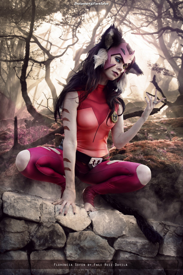 A female cosplayer dressed as a cat-like character with pink attire, posed on a rocky surface in a forest setting, surrounded by trees and ethereal lighting.