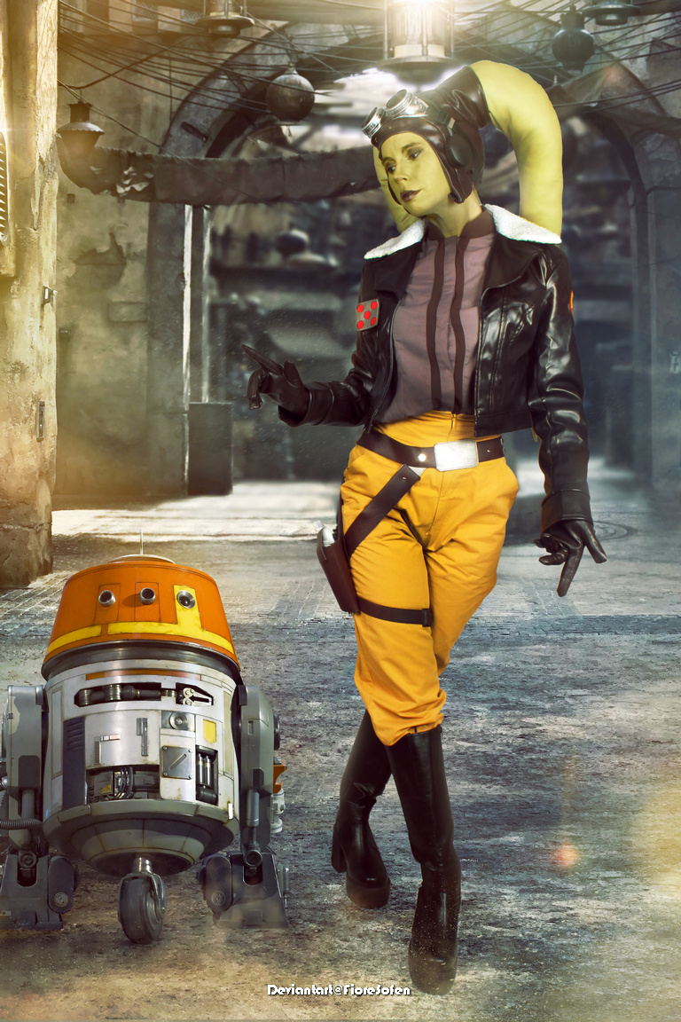 A character in a leather jacket and yellow pants, with head-tails and goggles, stands confidently next to a small, orange and gray droid in an industrial setting.