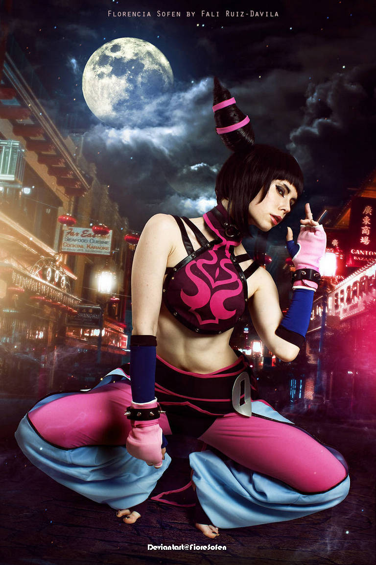 A female cosplayer dressed in a stylized outfit with vibrant pink and blue colors poses dramatically against a nighttime urban backdrop with a full moon.