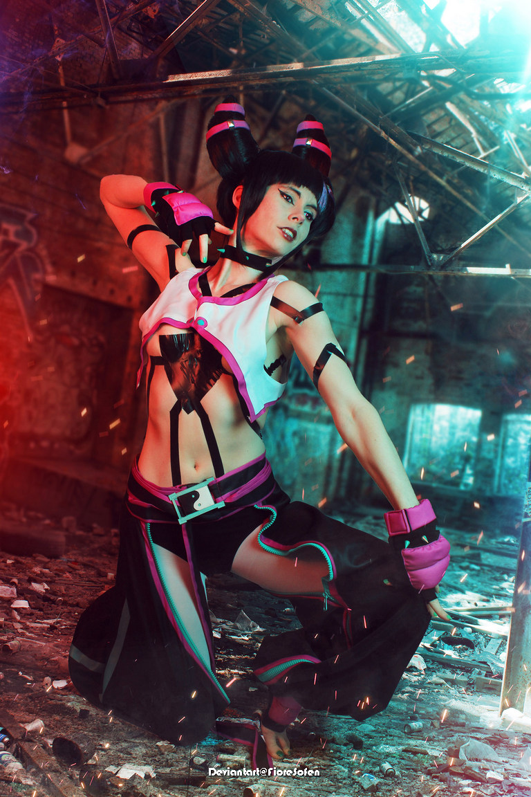A female cosplayer dressed in a futuristic outfit with pink and black accents striking a dynamic pose in an industrial setting with scattered debris and bright lighting.