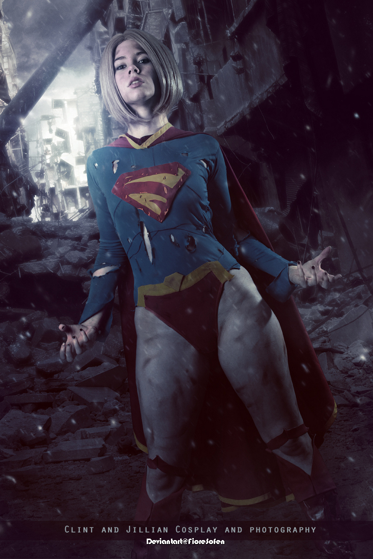 A female cosplayer dressed as Supergirl stands confidently amidst a post-apocalyptic backdrop, with a partially ruined cityscape and debris surrounding her.