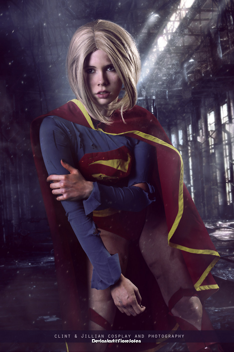 A young woman in a Supergirl costume poses dramatically in an abandoned building, with a soft light illuminating her features.