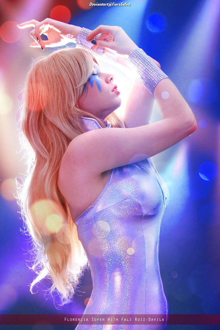 A model with long blonde hair poses gracefully, wearing a shimmering silver outfit and striking blue face paint, against a colorful blurred background.