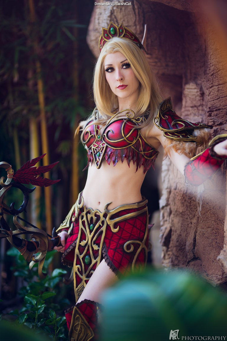 A cosplayer in elaborate red and gold armor poses confidently amidst greenery, showcasing a fantasy warrior character.