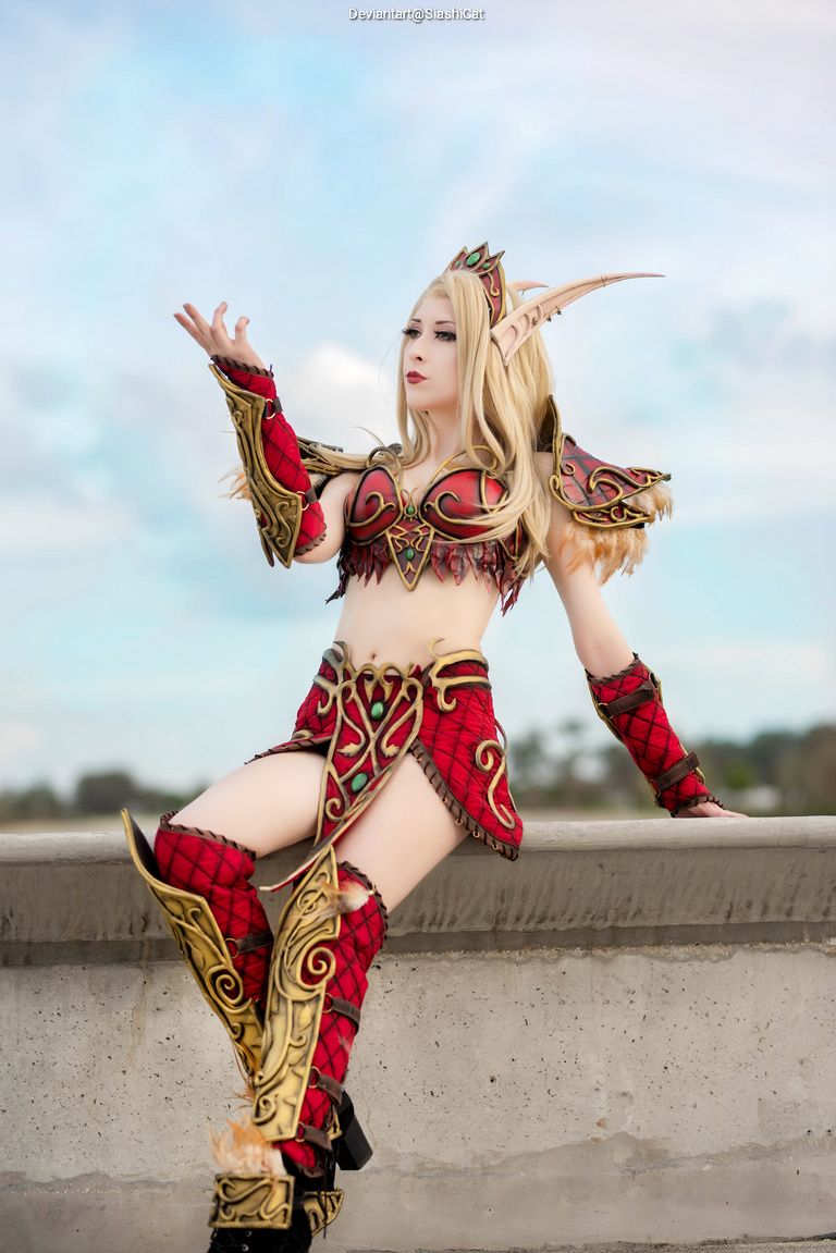 A female cosplayer wearing a detailed fantasy costume with red and gold accents, styled long blonde hair, and sharp ornamental shoulder pieces, posing confidently outdoors.