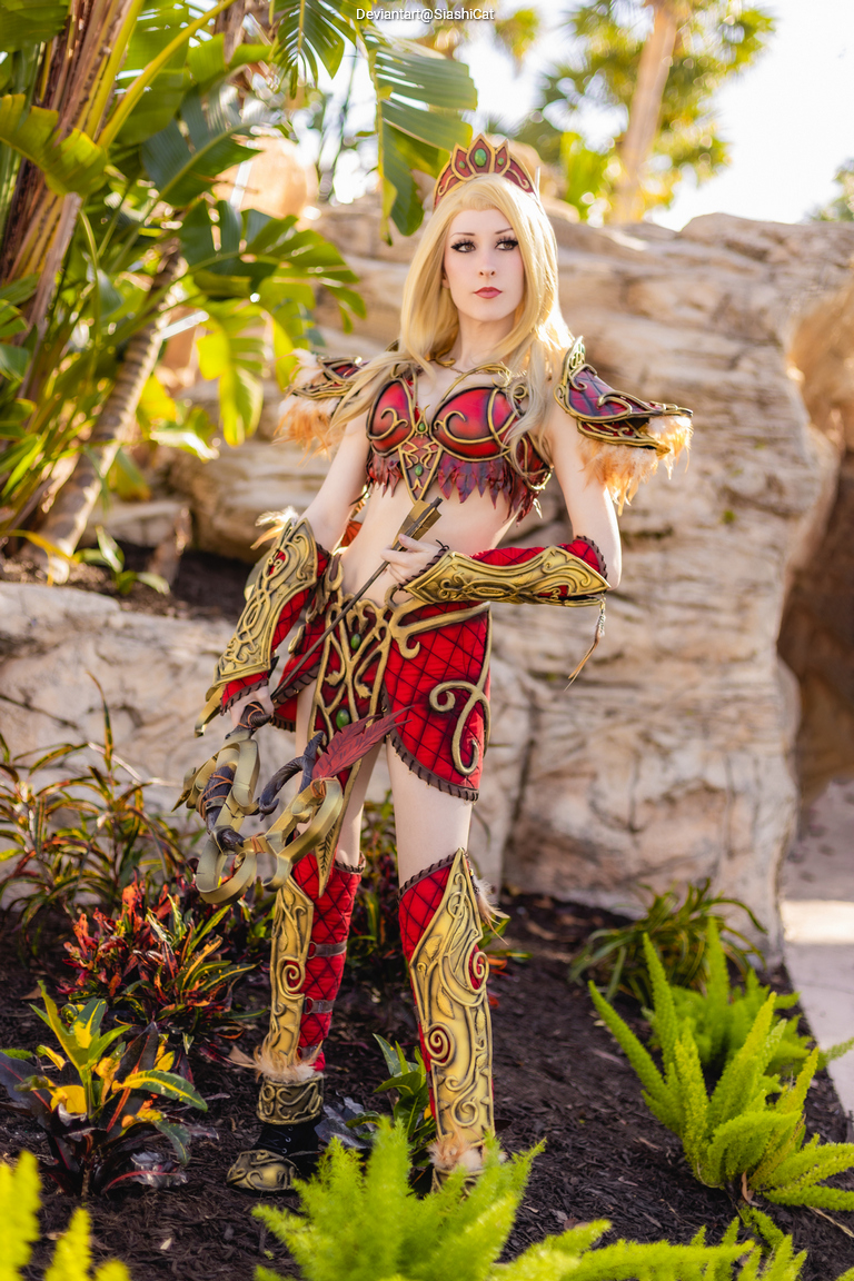 A person in a detailed fantasy costume with red and gold armor, posing confidently amidst lush greenery.