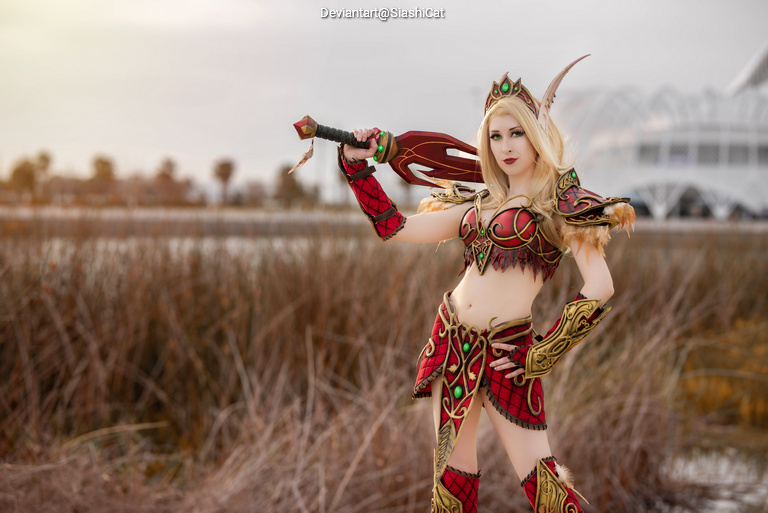 A female cosplayer dressed as a character with long blonde hair, wearing ornate red and green armor, holding a weapon, posed outdoors with a natural background.