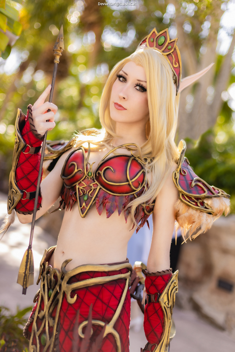 A person in a fantasy costume with red and gold armor, posing with a spear in a lush outdoor setting.
