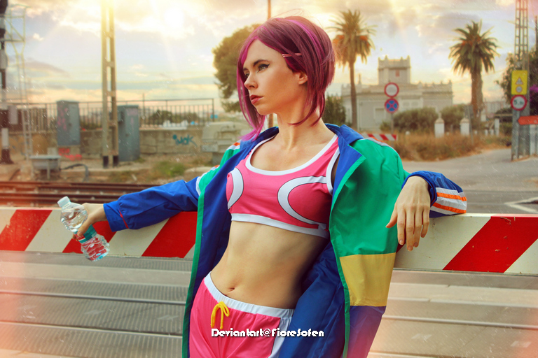 A person with pink hair poses confidently in a colorful athletic outfit, holding a water bottle, standing near a train crossing with palm trees in the background.