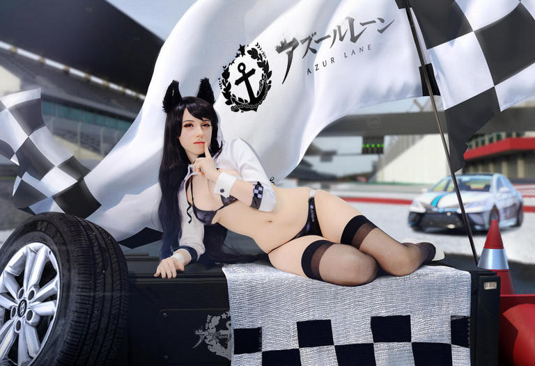 A female cosplayer dressed as a character from Azur Lane, lying on a checkered flag-themed racing-themed set with a car in the background.