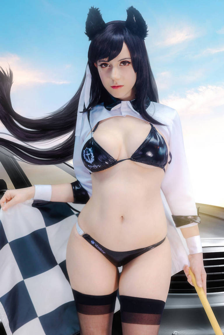 A woman dressed in a racing-themed outfit, featuring cat ears, a bikini top, and a checkered flag, poses confidently in front of a car against a blue sky backdrop.