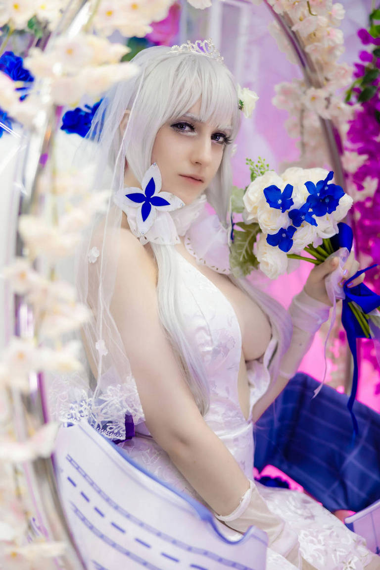 A person dressed in a white dress with long silver hair, posing with a bouquet of blue and white flowers, surrounded by floral decorations.