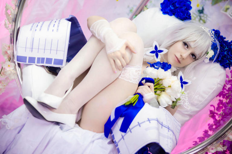 A person in a white dress and gloves sitting in a cozy chair, holding a bouquet of flowers, with a soft pink background accented by blue flowers.