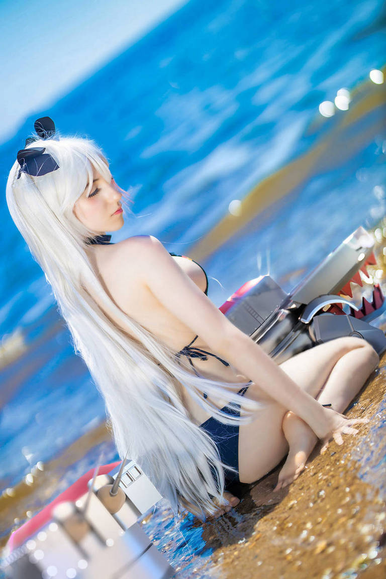 A person with long white hair sits on the beach in shallow water, wearing a black swimsuit and a black bow. They are surrounded by model figures resembling robotic creatures.