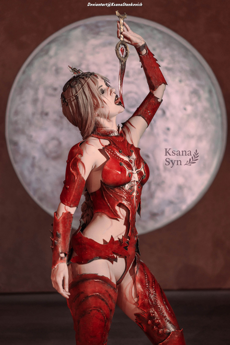 A woman in an intricate red costume with armor-like elements stands against a large moon backdrop, holding a decorative dagger to her mouth. Her makeup and hairstyle complement the dramatic theme.