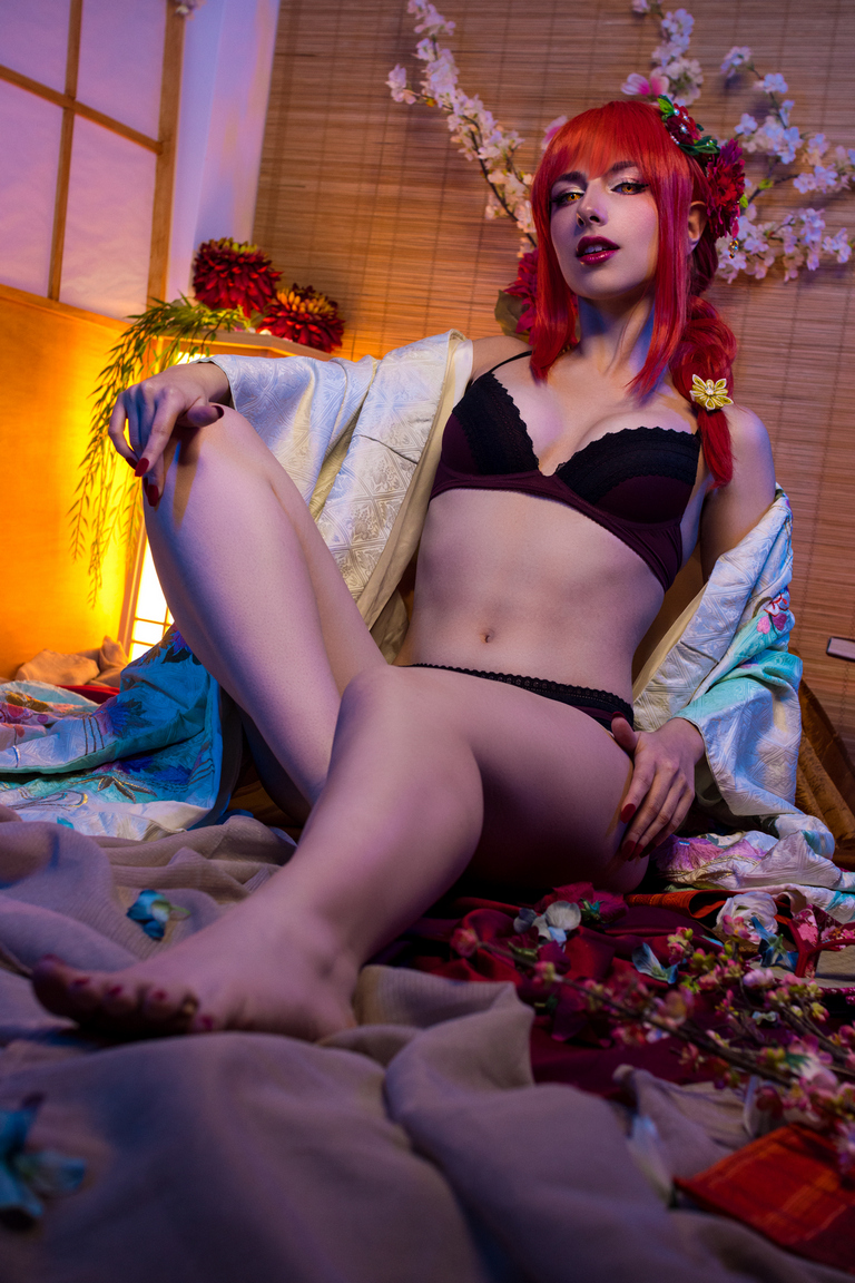 A person with red hair wearing a black bra and posing on a bed surrounded by flower petals and soft fabrics, with a wooden background.