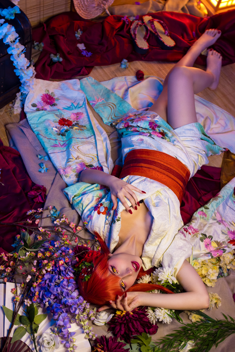A person with vibrant red hair lies on a bed of flowers, dressed in a colorful kimono. The setting is decorated with fabrics and accessories, creating a serene and artistic atmosphere.