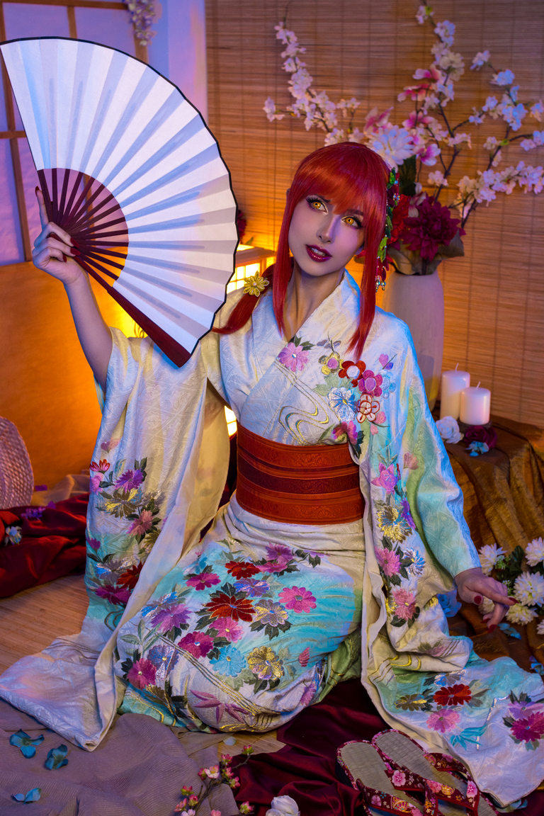 A person wearing a colorful kimono with floral embroidery, holding a traditional fan, surrounded by decorative flowers and candles.