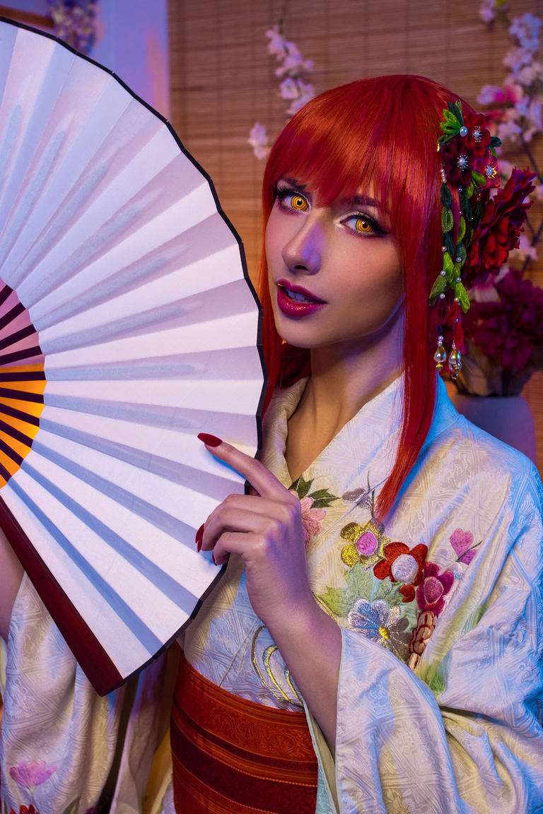A woman with red hair and striking yellow eyes poses holding a traditional fan, wearing a colorful kimono with floral patterns, set against a softly lit background.
