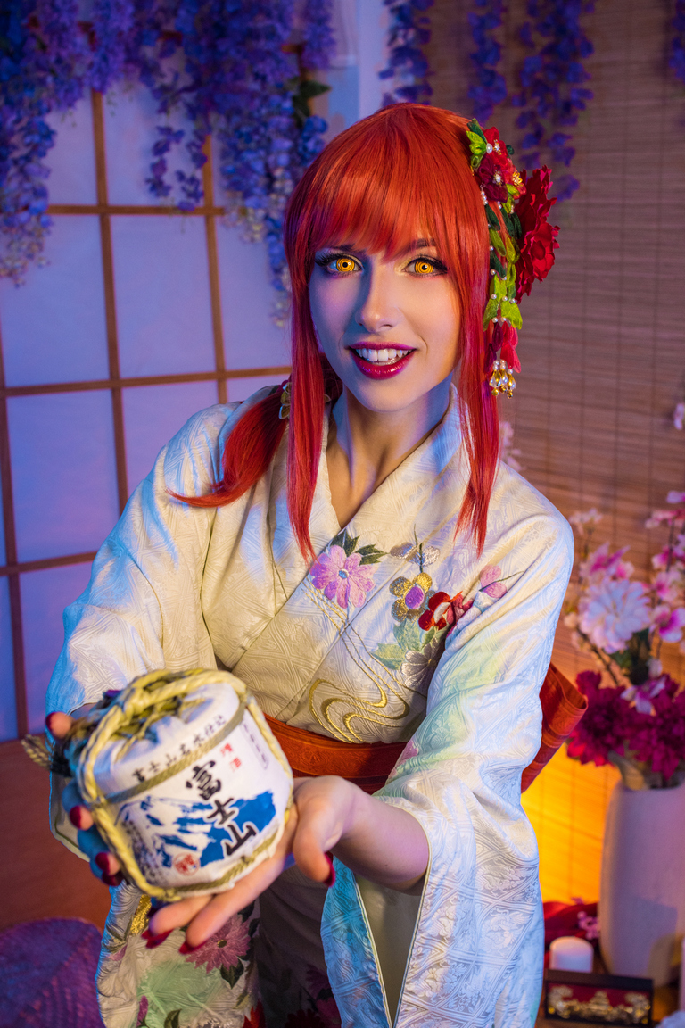 A person in traditional Japanese clothing (kimono) with vibrant red hair and yellow eyes is smiling while holding a decorative package, set against a floral and soft-lit background.
