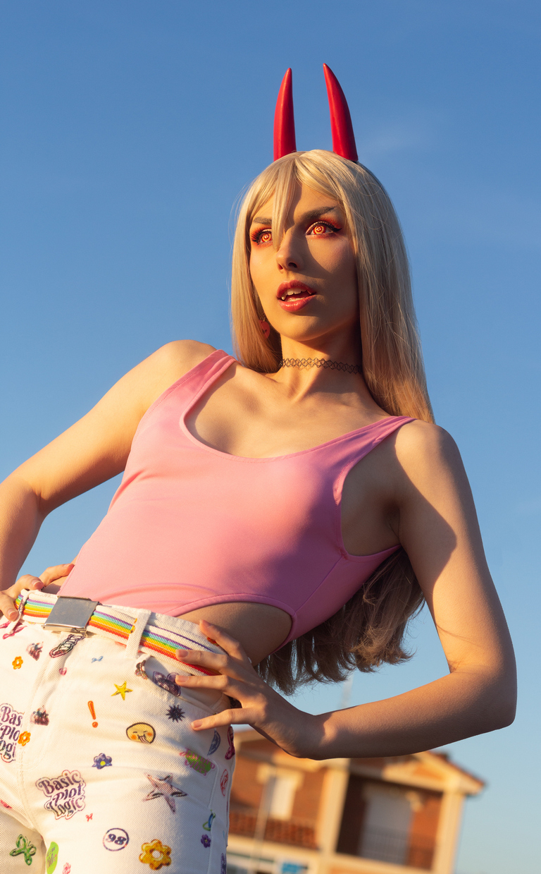 A person with long blonde hair and red horns poses outdoors, wearing a pink tank top and decorated white pants. The background features a clear blue sky.