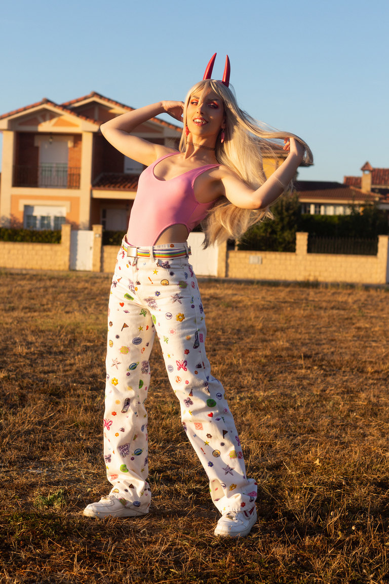 A young woman with long blonde hair and red horns is playfully posing outside, wearing a pink top and colorful patterned pants, against a sunny background.