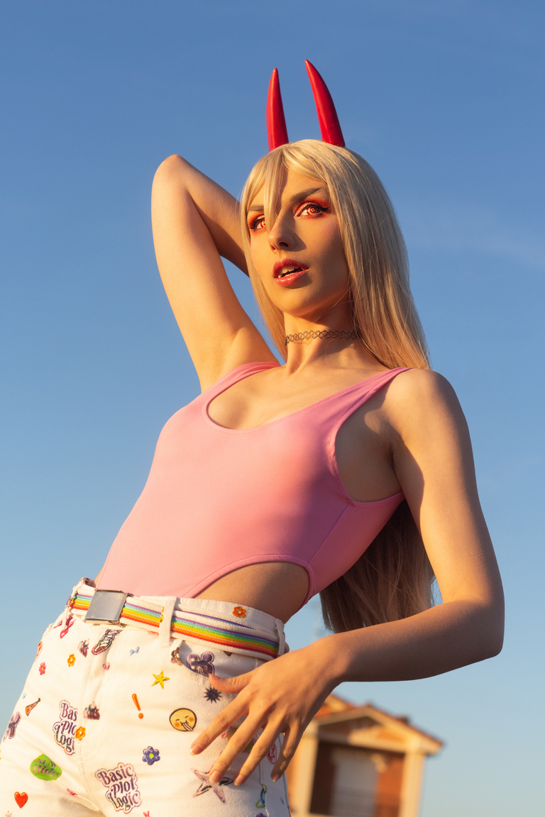 A person with long blonde hair, wearing a pink one-piece swimsuit and white shorts with colorful patterns, poses against a blue sky, sporting red devil horns and bold makeup.