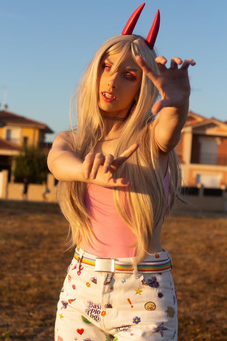 A person with long blonde hair and red devil horns poses outdoors, extending their hands towards the camera with a playful expression. They wear a pink top and colorful patterned pants, set against a sunset backdrop.