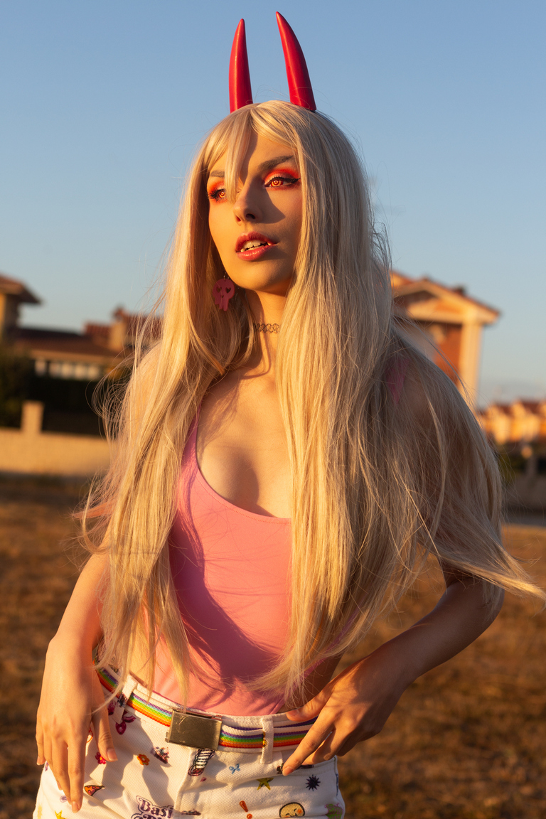 A person with long blonde hair and red horns stands against a sunset backdrop, wearing a pink top and patterned shorts. They have bold makeup with orange eyeshadow and accessories.
