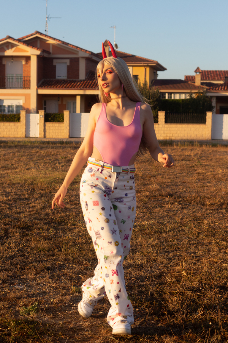 A person walking in a field during sunset, wearing a pink tank top and white pants with colorful patterns, featuring bunny ears and a soft expression.