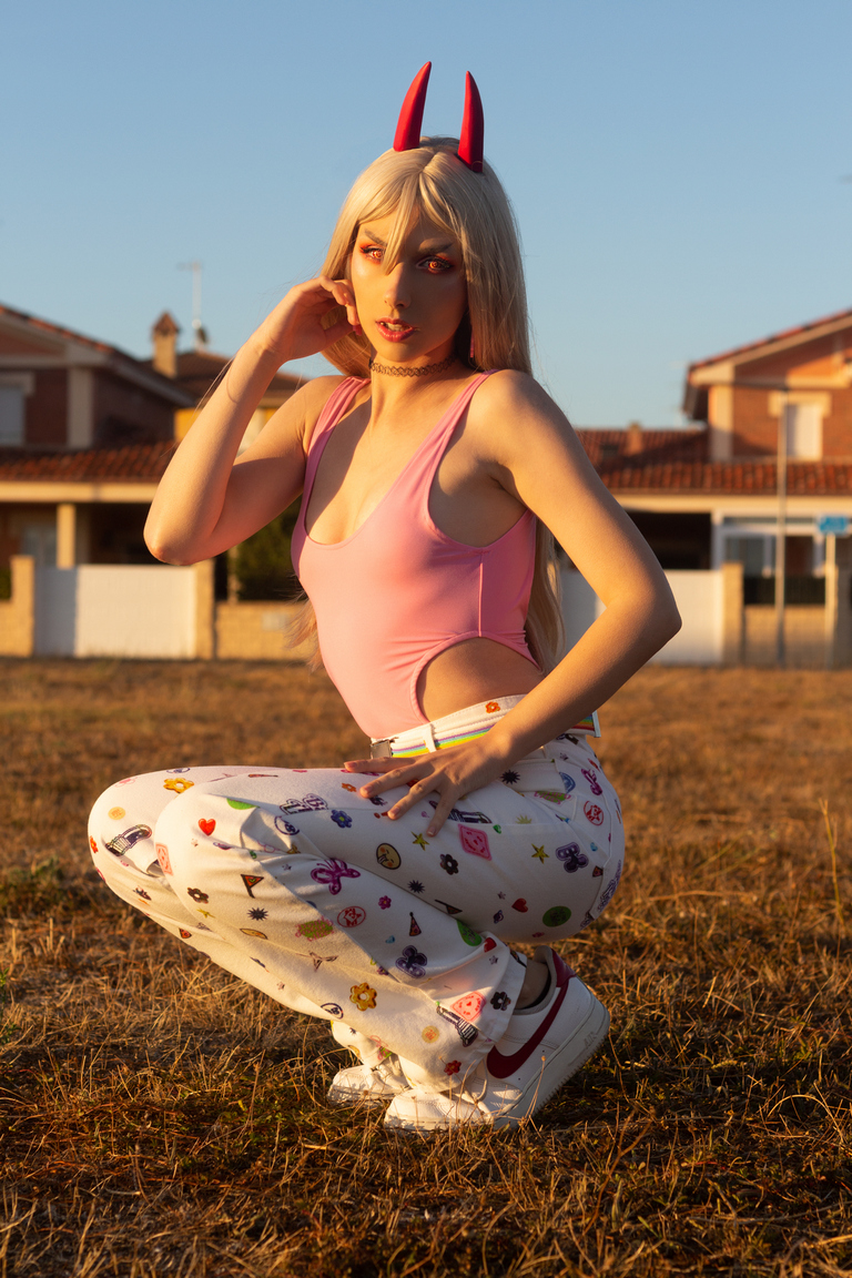 A person squatting in a field, wearing a pink tank top and white pants adorned with colorful patterns, with long blonde hair and red horns.