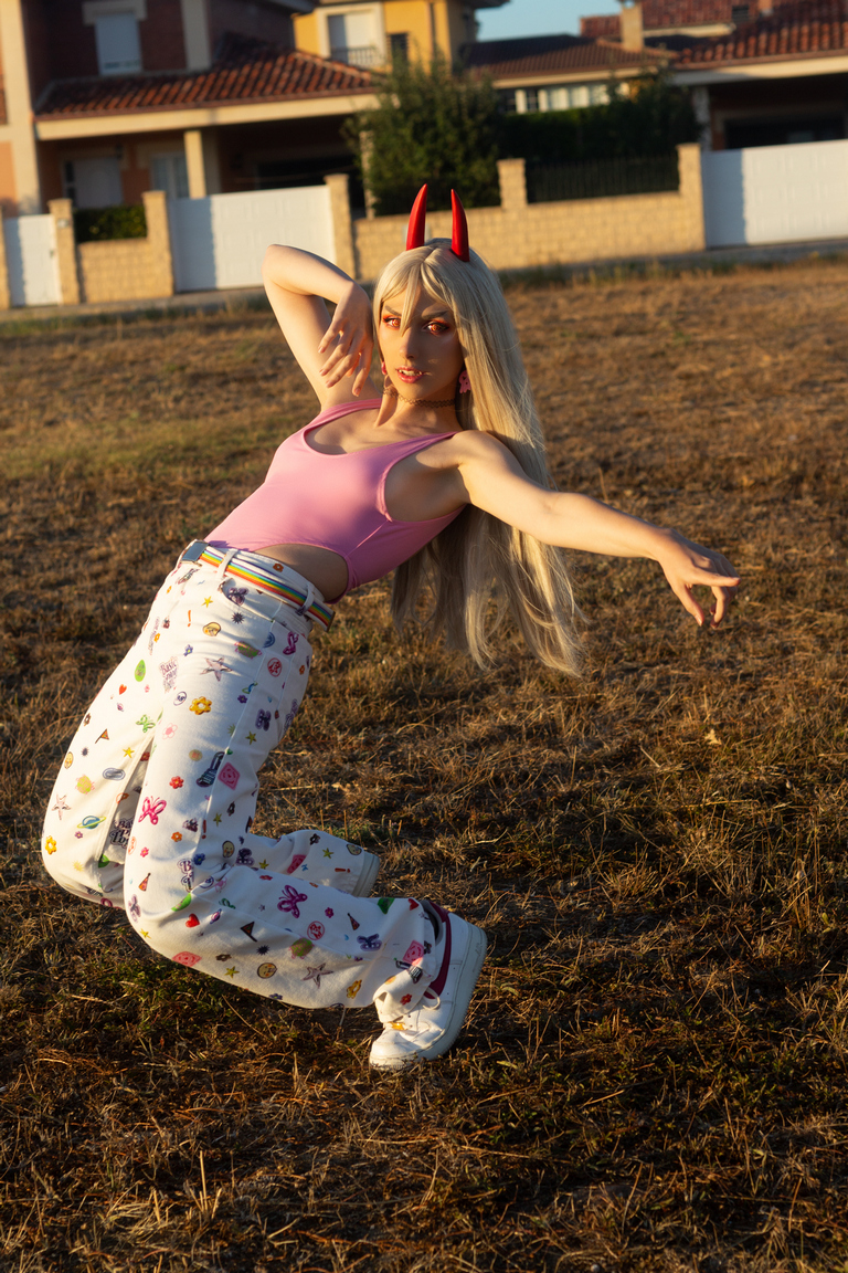 A person with long blonde hair and red horns wearing a pink top and patterned white pants poses playfully in a field during sunset.