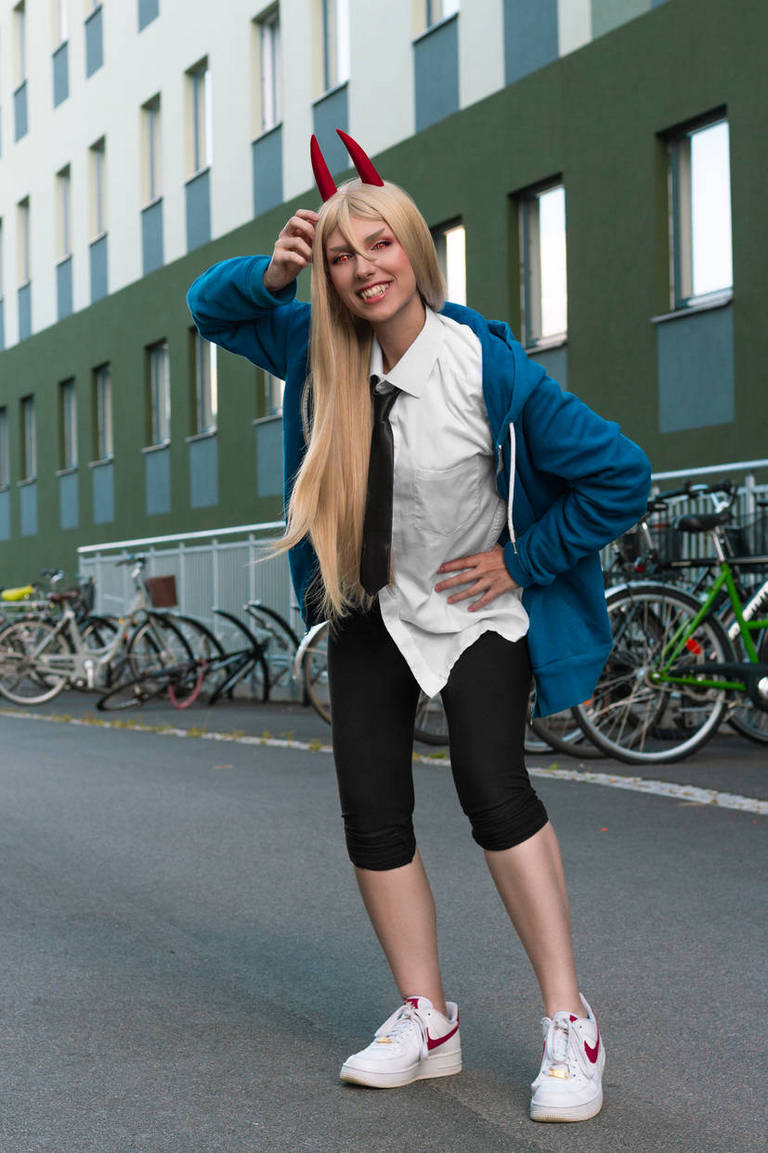 A person dressed in a casual outfit with a blue hoodie, white shirt, black pants, and red horns on their head, posing playfully outdoors with bicycles in the background.