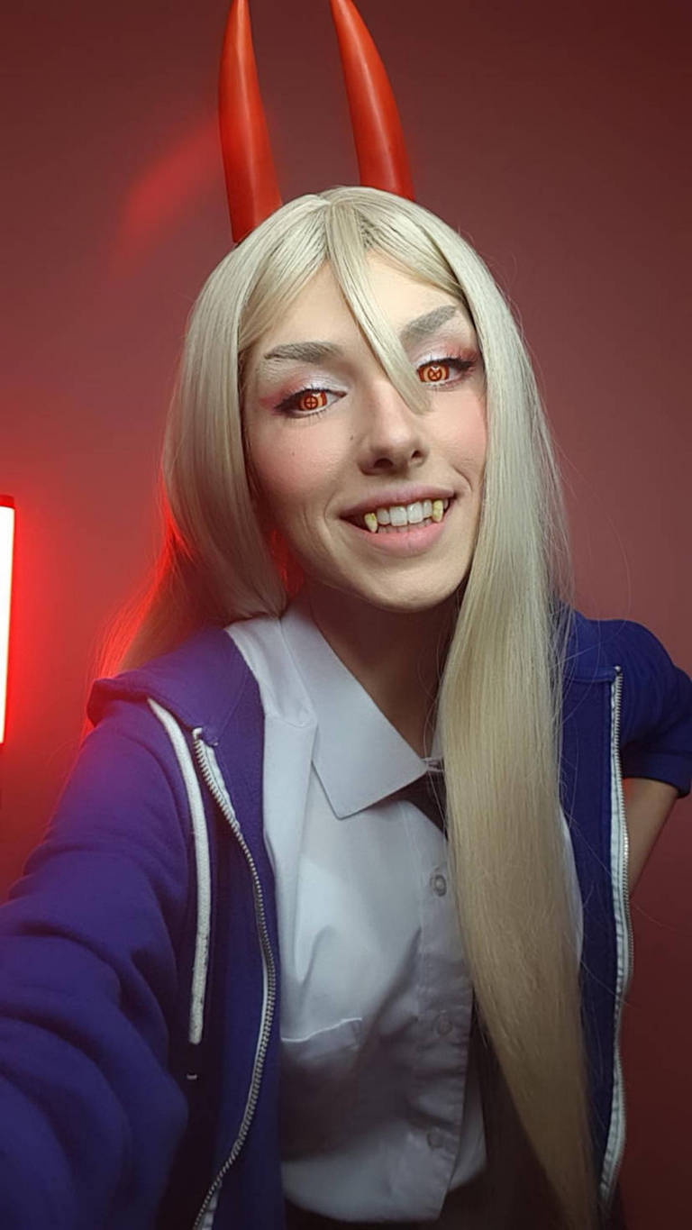 Person posing for a selfie with long blonde hair, red horns, and striking orange eyes, wearing a blue hoodie over a white shirt, against a red background.